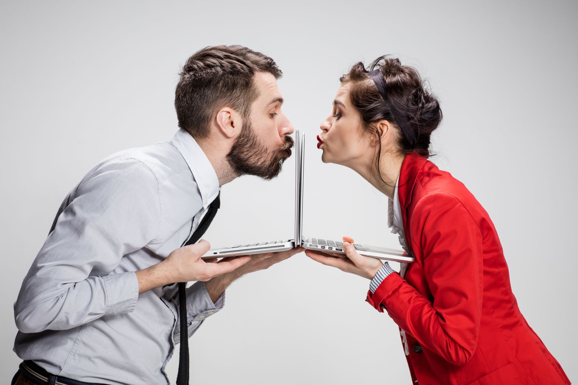 A man and a woman hold up their notebooks and each kisses their screen. The image depicts two singles meeting online who fall in love with each other.